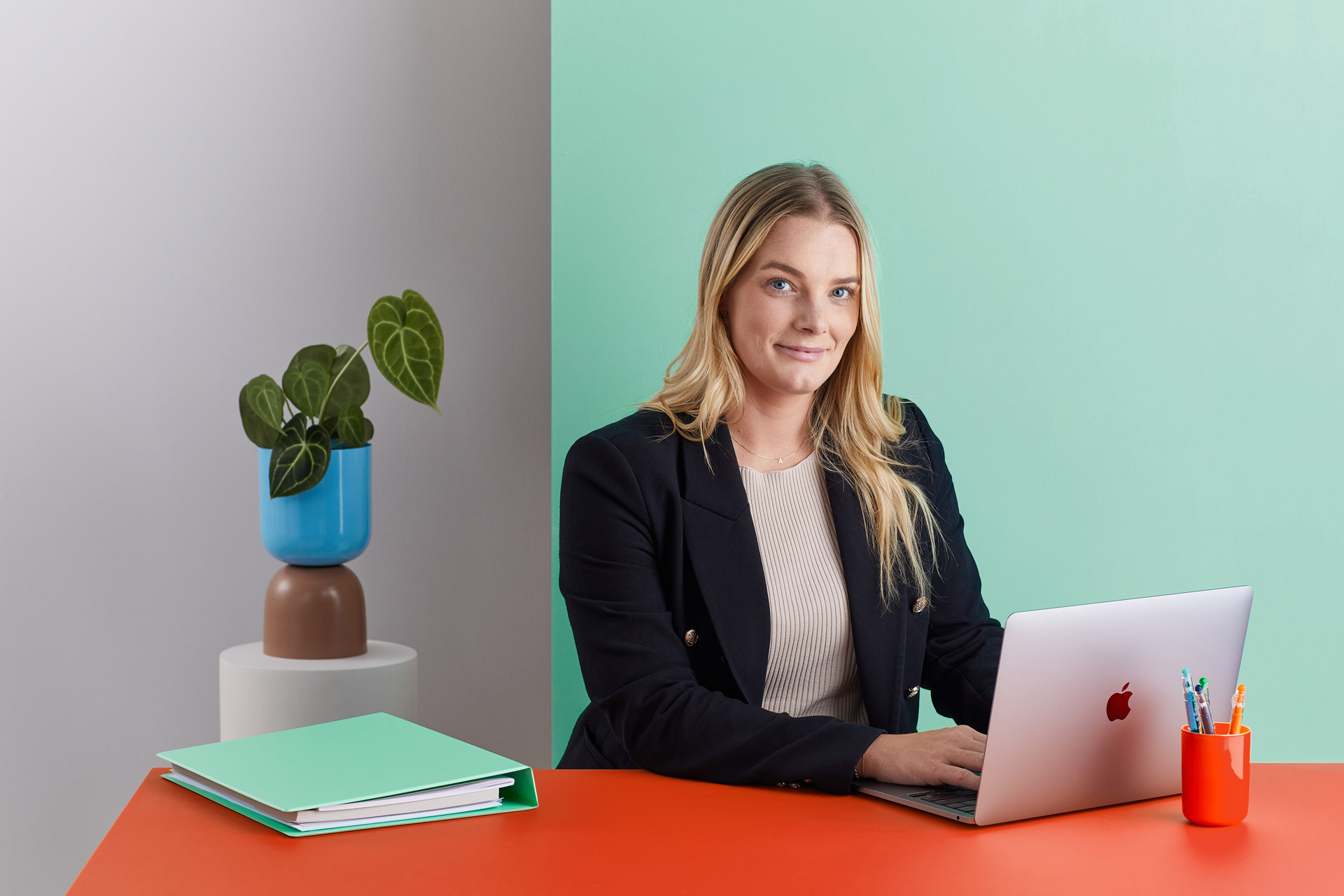 Amy Forwood, a legal professional, seated at a desk with a laptop, wearing a black blazer and a light top, with a green and red office setting in the background.